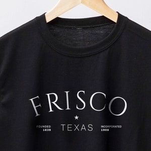 May include: Black t-shirt with the word "FRISCO" in a curved font, with "TEXAS" and the founding and incorporation dates below. The shirt is hanging on a wooden hanger against a white background.