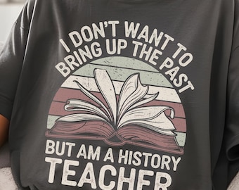 Puede incluir: Camiseta gris oscuro con un gráfico que presenta un libro abierto y el texto "I DON'T WANT TO BRING UP THE PAST BUT AM A HISTORY TEACHER" en blanco. El diseño incluye una puesta de sol de estilo retro con rayas rojas, rosas y verdes.