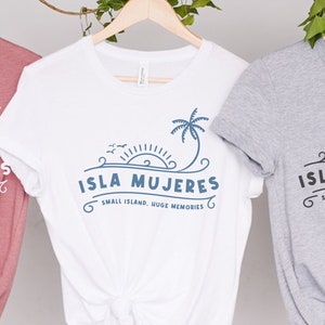 May include: Three t-shirts in different colors: dusty rose, white, and gray. Each shirt features the text "ISLA MUJERES" with a palm tree and sun design. The phrase "SMALL ISLAND, HUGE MEMORIES" is below the main text.