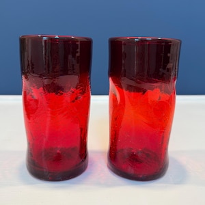 May include: Two cylindrical drinking glasses made of vibrant red glass with a distinct crackle texture throughout the body. The glasses feature a smooth, solid red rim at the top and a slightly irregular, organic shape with a thick, weighted base.