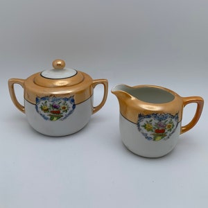 May include: A vintage porcelain creamer and sugar bowl set with gold accents and floral designs. The creamer and sugar bowl are white with a gold band around the top and a floral design in the center.
