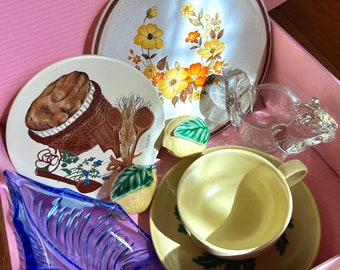 May include: A collection of vintage kitchenware including two ceramic plates with floral and basket motifs, a yellow teacup and saucer, a blue glass leaf-shaped dish, a clear glass creamer, and two small ceramic lemon-shaped decorative items.