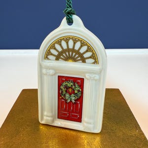 May include: A white ceramic Christmas ornament shaped like a doorway. The door is red with a green wreath and bow. Above the door is a gold and yellow stained glass window design. A green cord is attached for hanging.