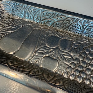 May include: A rectangular silver-toned metal tray featuring an embossed design of various fruits including pears, apples, and grapes, surrounded by a border of textured leaves and berries. The metallic surface has a polished finish.