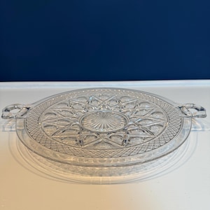 May include: A clear glass serving tray with a starburst design in the center and a diamond pattern around the edge. The tray has two handles and is sitting on a white surface. The background is a dark blue.