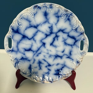 May include: A round ceramic serving plate featuring a blue and white mottled pattern with a crackle-like effect. The plate has two integrated side handles and a scalloped, embossed rim with subtle gold-toned accents along the edges.