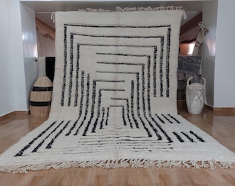May include: A white and black geometric patterned rug with a fringe border. The rug has a modern design with a series of black lines creating a geometric pattern on a white background.