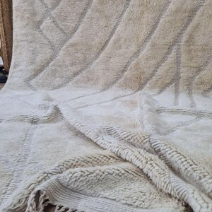 K&ouml;nnte beinhalten: Beige-wei&szlig;er marokkanischer Teppich mit geometrischem Design. Der Teppich hat diagonale Linien und eine strukturierte Oberfl&auml;che. Der untere Rand hat Fransen. Der Teppich besteht aus einem weichen Material.