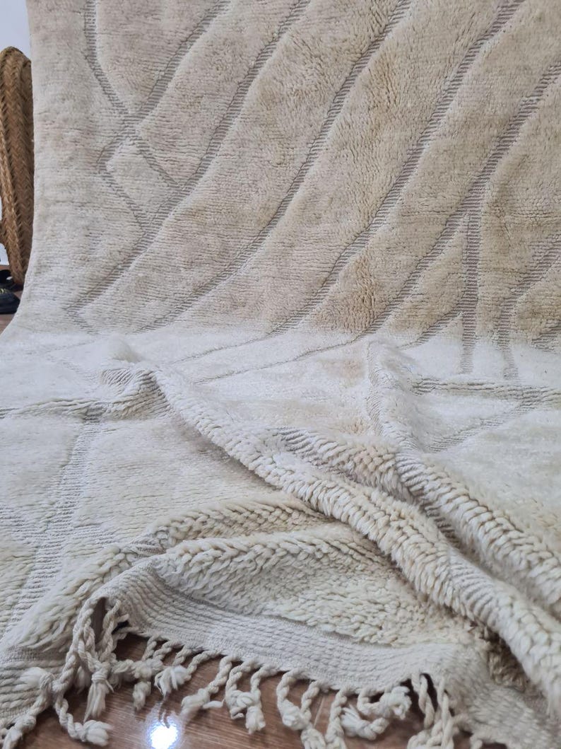 K&ouml;nnte beinhalten: Beige-wei&szlig;er marokkanischer Teppich mit geometrischem Design. Der Teppich hat diagonale Linien und eine strukturierte Oberfl&auml;che. Der untere Rand hat Fransen. Der Teppich besteht aus einem weichen Material.