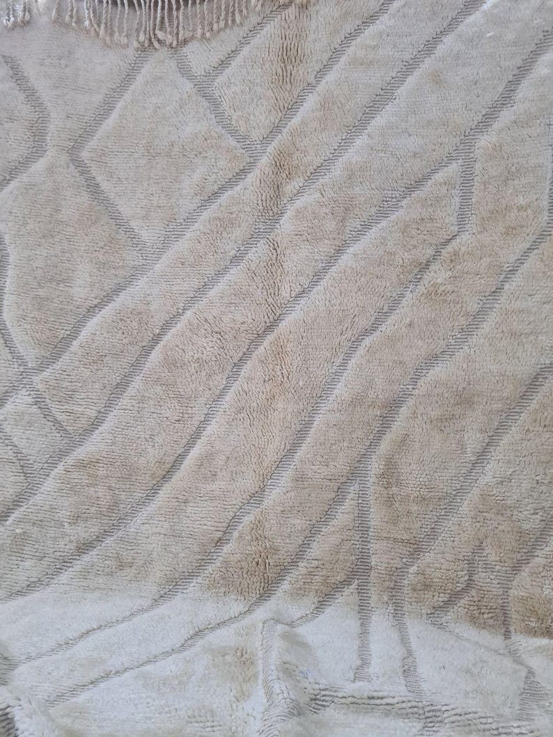 K&ouml;nnte beinhalten: Beiger Teppich mit geometrischem Muster. Der Teppich hat diagonale Streifen und Rauten in einem helleren Beigeton. Der Teppich hat eine strukturierte Oberfl&auml;che und einen Fransenbesatz oben.
