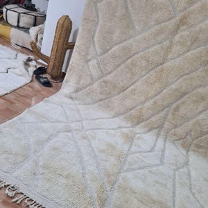 K&ouml;nnte beinhalten: Ein gro&szlig;er, beigefarbener Teppich mit geometrischen Mustern und einem Fransenrand. Der Teppich hat eine strukturierte Oberfl&auml;che mit Diamant- und linearen Designs in einem helleren Beigeton. Der Teppich wird auf einem Holzboden pr&auml;sentiert.