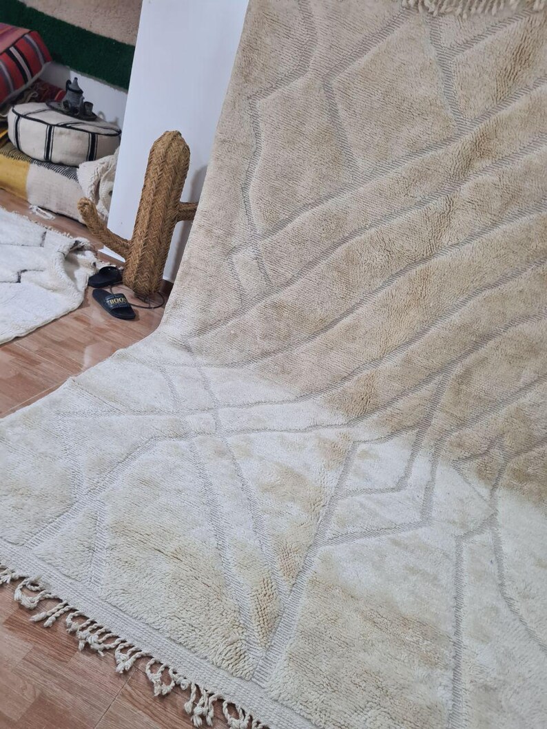 K&ouml;nnte beinhalten: Ein gro&szlig;er, beigefarbener Teppich mit geometrischen Mustern und einem Fransenrand. Der Teppich hat eine strukturierte Oberfl&auml;che mit Diamant- und linearen Designs in einem helleren Beigeton. Der Teppich wird auf einem Holzboden pr&auml;sentiert.