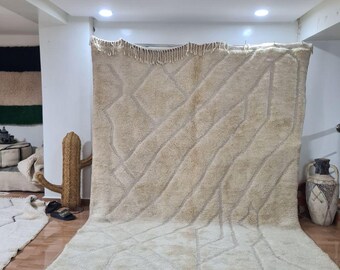 May include: A large, beige Moroccan rug with a geometric diamond pattern. The rug has a thick pile and a fringe along the bottom edge. The rug is displayed indoors, with a cactus sculpture and a decorative vase in the background.