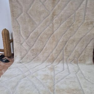 K&ouml;nnte beinhalten: Ein gro&szlig;er, beigefarbener Teppich mit einem geometrischen Muster aus Rauten und diagonalen Linien. Der Teppich hat einen dicken, strukturierten Flor und einen Fransenrand. Das Design ist in einem helleren Beigeton gehalten, was einen subtilen Kontrast erzeugt.