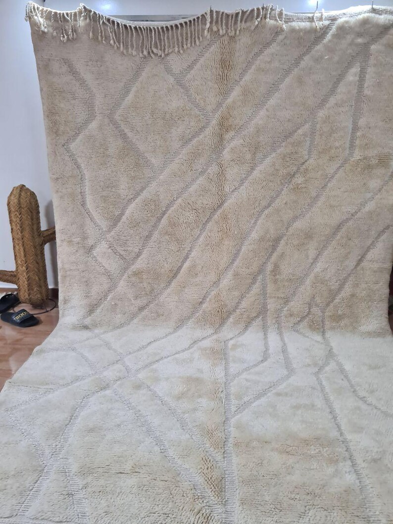 K&ouml;nnte beinhalten: Ein gro&szlig;er, beigefarbener Teppich mit einem geometrischen Muster aus Rauten und diagonalen Linien. Der Teppich hat einen dicken, strukturierten Flor und einen Fransenrand. Das Design ist in einem helleren Beigeton gehalten, was einen subtilen Kontrast erzeugt.