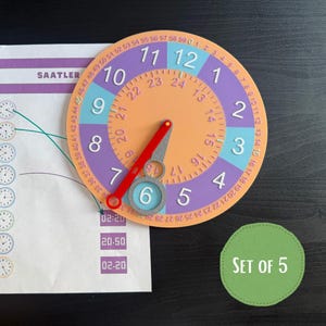 Op de afbeelding: Een rond educatief klokinstrument met een perzikkleurig midden, paarse en blauwe segmenten en witte cijfers van 1 tot 12. Het heeft een beweegbare rode en grijze wijzer, een buitenste ring met minuten 0 tot 59 en een binnenring met uren 13 tot 24.