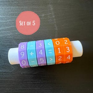 Pode incluir: Uma ferramenta de manipulação matemática com uma haste cilíndrica branca e cinco anéis hexagonais rotativos em roxo, azul e laranja. Os anéis exibem números e símbolos matemáticos brancos. Um círculo rosa contém o texto SET OF 5.