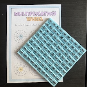 May include: A light blue square silicone tray featuring a grid of multiplication equations from 1x1 to 10x10, placed over a white paper worksheet titled Multiplication Wheel with circular diagrams for mental math practice.