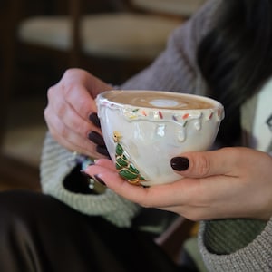 May include: A white ceramic coffee cup with a Christmas tree design. The cup has a glossy finish with colorful accents around the rim. The tree is green with gold accents. The cup is filled with a light brown beverage.