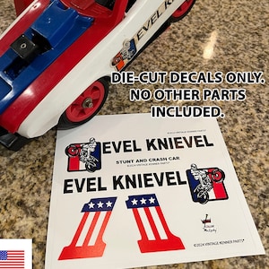 May include: Die-cut decals for a vintage Evel Knievel Stunt and Crash Car toy. The decals are white with red, blue, and black accents. The decals feature the words "Evel Knievel" and a star-spangled banner design.
