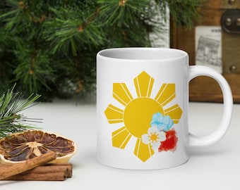 Puede incluir: Taza de cerámica blanca con un sol amarillo y ocho rayos, que representa la bandera filipina, con tres flores de acuarela en la esquina inferior derecha.
