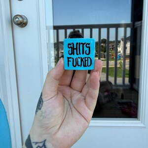 May include: A small, square, bright turquoise block featuring the black text "SHITS FUCKED" printed in a bold, stylized font on the front face. The object has a smooth, solid appearance and is held up against a glass door background.