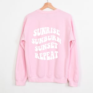 Peut inclure: Sweat-shirt rose à manches longues avec un texte blanc au dos qui dit "Sunrise Sunburn Sunset Repeat" dans une police rétro.