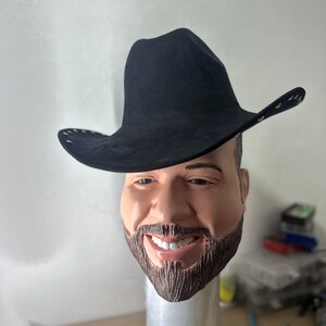 May include: A black cowboy hat with a wide brim and a stitched detail around the edge. The hat is worn by a person with a beard and a smiling face.
