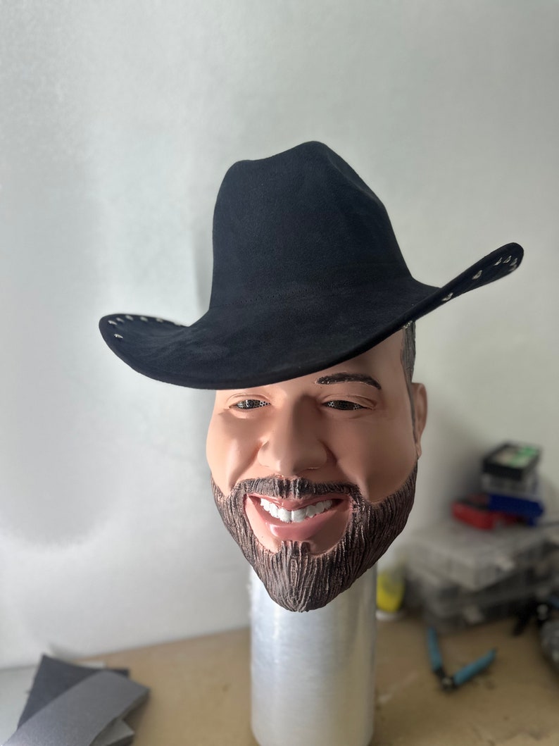 May include: A black cowboy hat with a wide brim and a stitched detail around the edge. The hat is worn by a person with a beard and a smiling face.