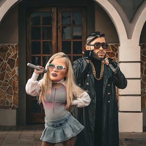 May include: A digitally altered image of two figures, one a blonde woman in a denim skirt and the other a man in a black leather coat, both holding microphones. The woman wears sunglasses and a pink and gray bodysuit. The man wears a gold chain.
