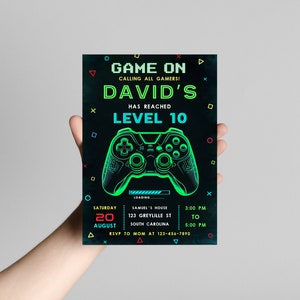 May include: A black and green video game party invitation with a neon green and blue gamepad graphic. The invitation reads "Game On Calling All Gamers! David's Has Reached Level 10". The invitation includes the date, time, and location of the party.