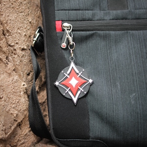 K&ouml;nnte beinhalten: Ein schwarzer Rucksack mit einem roten Rei&szlig;verschluss und einem silbernen Schl&uuml;sselanh&auml;nger mit einem schwarzen und wei&szlig;en sternf&ouml;rmigen Anh&auml;nger. Der Stern hat eine rote Mitte.