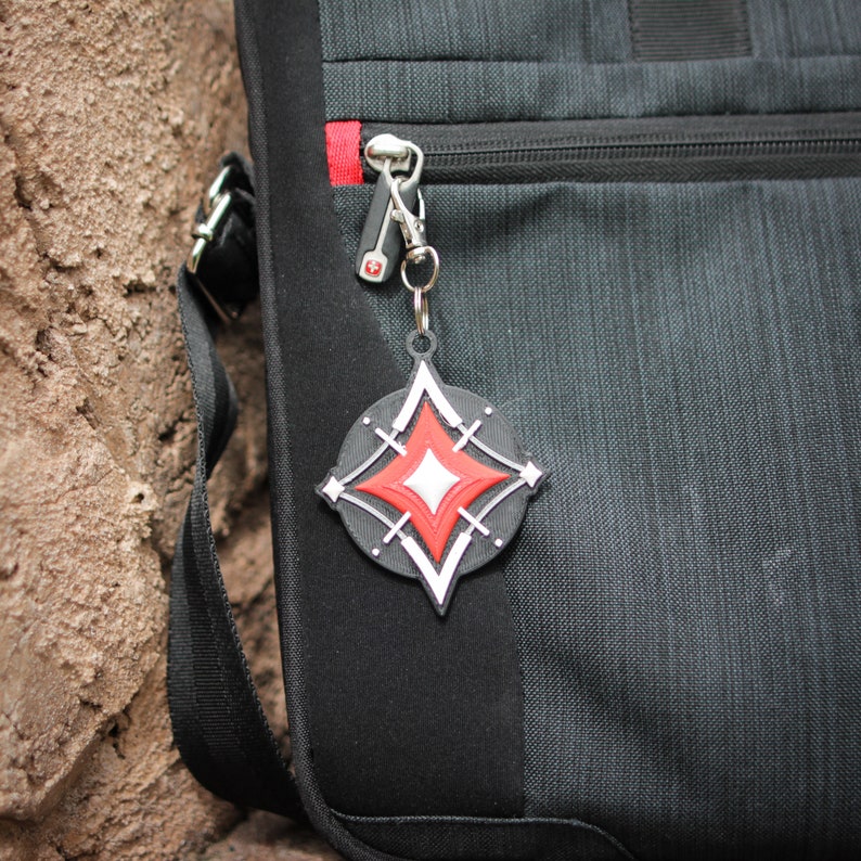 K&ouml;nnte beinhalten: Ein schwarzer Rucksack mit einem roten Rei&szlig;verschluss und einem silbernen Schl&uuml;sselanh&auml;nger mit einem schwarzen und wei&szlig;en sternf&ouml;rmigen Anh&auml;nger. Der Stern hat eine rote Mitte.