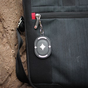 K&ouml;nnte beinhalten: Eine schwarze Tasche mit einem silbernen Rei&szlig;verschluss und einem silbernen und schwarzen Schl&uuml;sselanh&auml;nger mit einer Diamantenform in der Mitte.