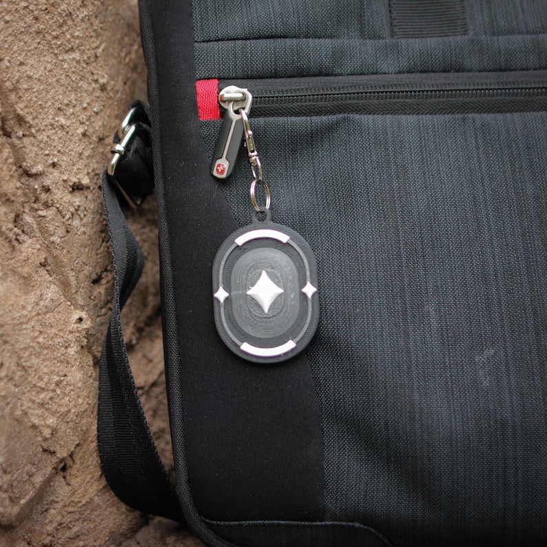 K&ouml;nnte beinhalten: Eine schwarze Tasche mit einem silbernen Rei&szlig;verschluss und einem silbernen und schwarzen Schl&uuml;sselanh&auml;nger mit einer Diamantenform in der Mitte.