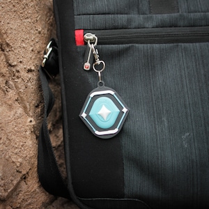K&ouml;nnte beinhalten: Eine schwarze Tasche mit einer Rei&szlig;verschlusstasche und einem silbernen Schl&uuml;sselanh&auml;nger mit einem blauen und wei&szlig;en geometrischen Anh&auml;nger.