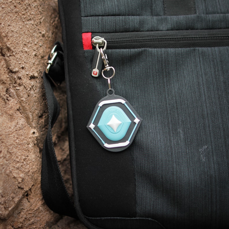 K&ouml;nnte beinhalten: Eine schwarze Tasche mit einer Rei&szlig;verschlusstasche und einem silbernen Schl&uuml;sselanh&auml;nger mit einem blauen und wei&szlig;en geometrischen Anh&auml;nger.