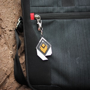 K&ouml;nnte beinhalten: Eine schwarze und graue Laptoptasche mit einem silbernen Rei&szlig;verschluss und einem wei&szlig;en und goldenen Schl&uuml;sselanh&auml;nger mit geometrischem Design.