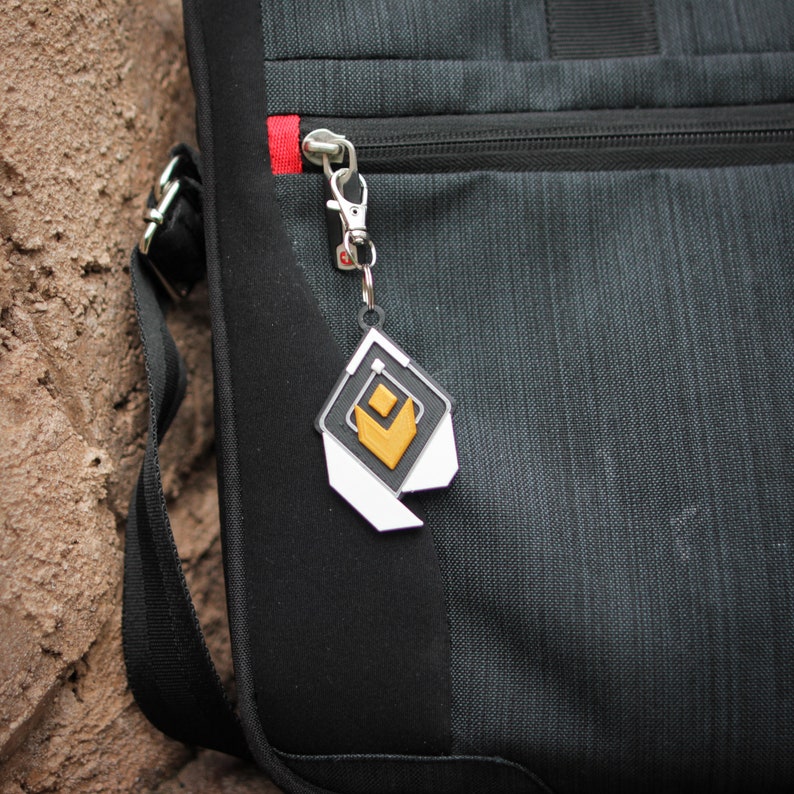 K&ouml;nnte beinhalten: Eine schwarze und graue Laptoptasche mit einem silbernen Rei&szlig;verschluss und einem wei&szlig;en und goldenen Schl&uuml;sselanh&auml;nger mit geometrischem Design.