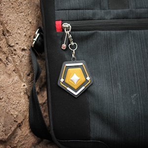 K&ouml;nnte beinhalten: Ein schwarzer und grauer Rucksack mit einem silbernen Rei&szlig;verschluss und einem goldenen und wei&szlig;en Schl&uuml;sselanh&auml;nger.