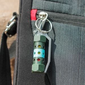 May include: A green and blue keychain shaped like a grenade with a silver ring and clip.
