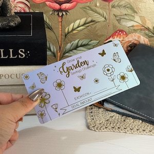 May include: A rectangular savings challenge card with a light blue gradient background. The card features gold foil illustrations of flowers and butterflies, with the text "Grow your Garden Savings Challenge," "EACH ICON =," and "GOAL AMOUNT."