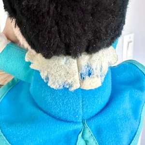 May include: A blue and white plush toy with a black fuzzy head. The toy has a white collar with some wear and tear.