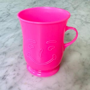 May include: A vibrant pink plastic mug featuring a smiling face design and a handle. The mug has a wide opening and a small base. The mug is made of a solid, opaque plastic material.