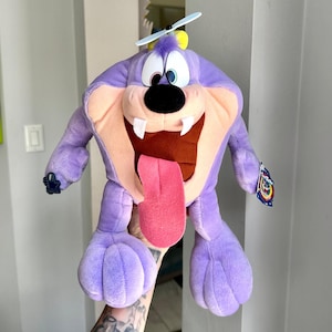 May include: A purple plush toy character with a wide open mouth, two white teeth, and a long pink tongue. The character features large white eyes, a black nose, and a small blue propeller on its head. A small tag is attached to the left arm.