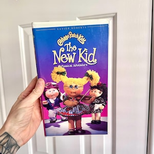 May include: A VHS tape of the Cabbage Patch Kids movie "The New Kid: A Musical Adventure". The cover features three Cabbage Patch Kids dolls, one playing the violin, one singing, and one dancing. The background is purple.