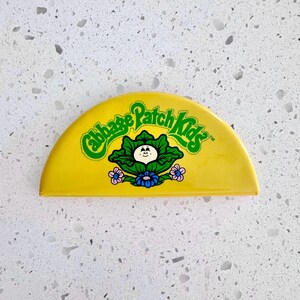May include: A yellow, semi-circular Cabbage Patch Kids pin. The pin features the Cabbage Patch Kids logo in green, along with a graphic of a cabbage with a face, blue and pink flowers.