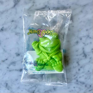 May include: A green Kermit the Frog-shaped magic sponge in a clear plastic package. The package has the text "Jim Henson's Muppet Babies" and "Baby Kermit Magic Sponge Fragranced Soap Filled Sponge". The net weight is .75 oz.