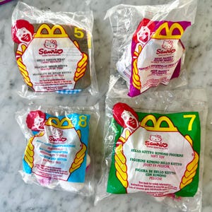 May include: Four sealed McDonald's Happy Meal toys featuring Hello Kitty and Dear Daniel characters. Each package has a different number and a Ronald McDonald graphic. The toys include a fabric bracelet, stamp mobile toy, hair scrunchy, and kimono figurine.
