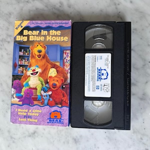 May include: A VHS tape and its cover for "Bear in the Big Blue House." The cover features a large brown bear with other colorful characters. The tape is black with a white label.